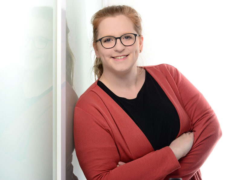 A portrait image of Elisa Mink. She leans against a glass wall and smiles friendly.